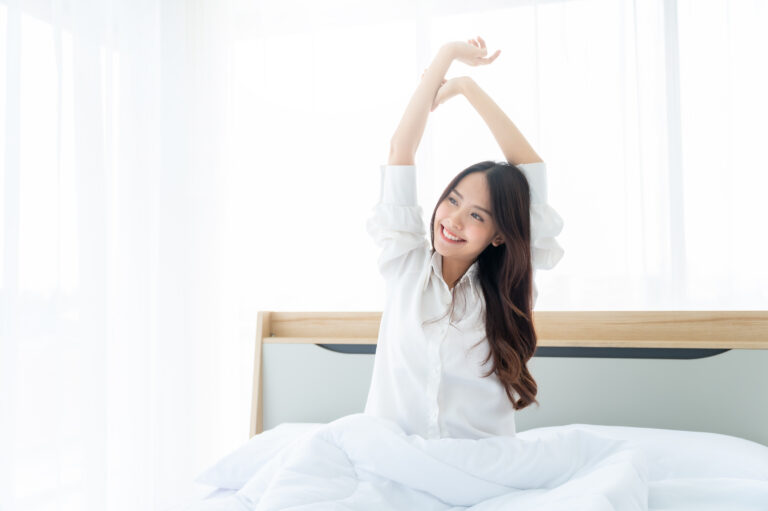 Woman sitting up in bed stretching her arms overhead in front of a sunlit window
