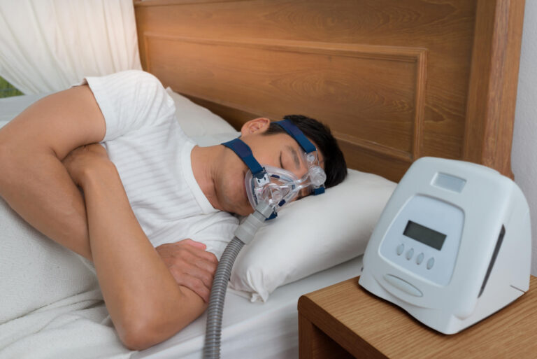 Man asleep on his side wearing a CPAP mask, the machine running on the nightstand beside him