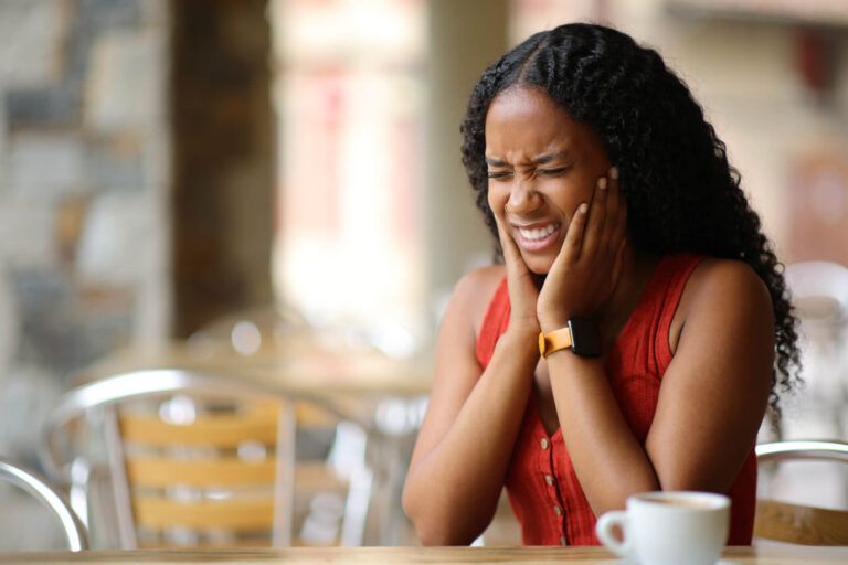 Woman at a cafe table wincing and holding her jaw with both hands