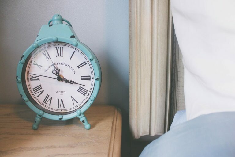 Teal retro alarm clock sitting on a wooden nightstand beside a bed