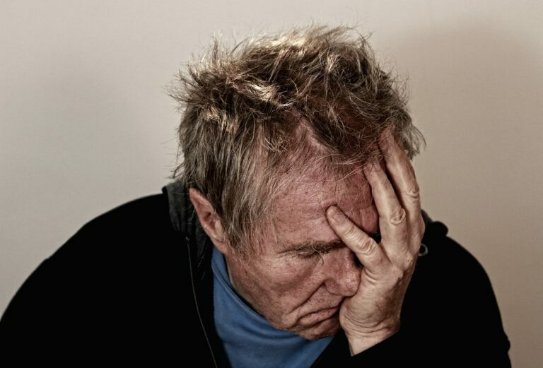 Grey-haired man in a dark top covering his face with one hand, looking worn out