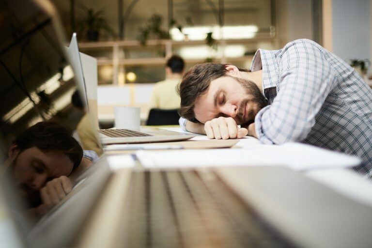 Businessman slumped asleep on his laptop at an open-plan office desk