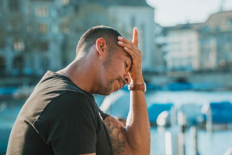 Man in a dark T-shirt pressing one hand to his forehead beside a waterfront