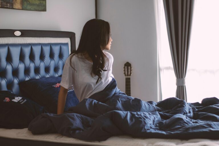 Woman sitting on the edge of an unmade bed in a dim bedroom, seen from behind