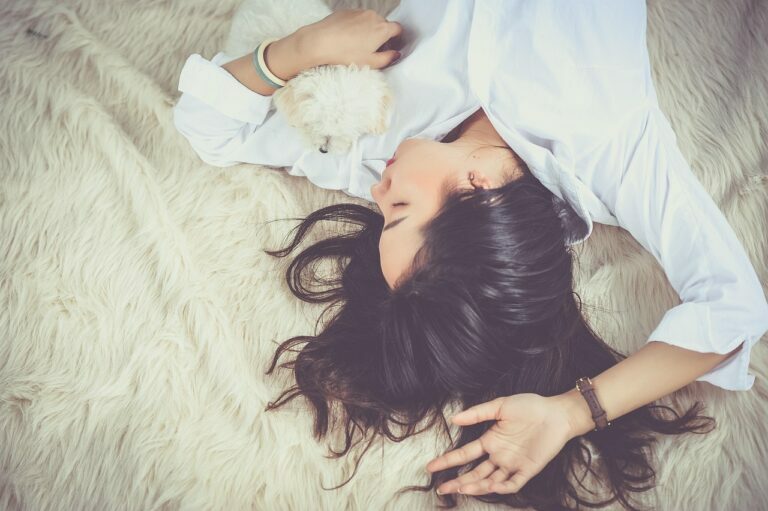 Woman lying asleep on a soft white blanket in calm natural light, a small dog curled beside her